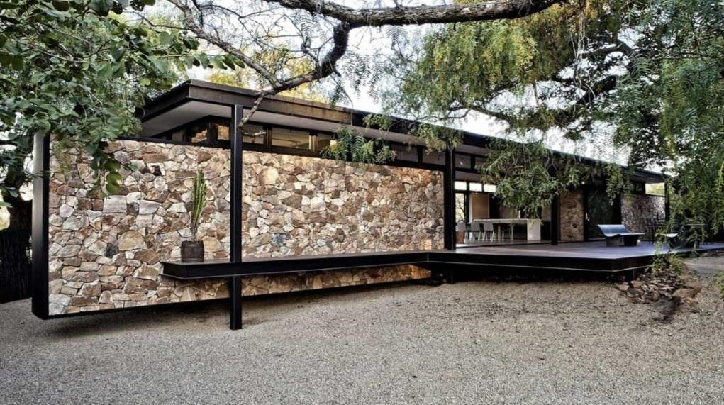 A long stone and black-steel house under trees, with a timber deck and gravel court
