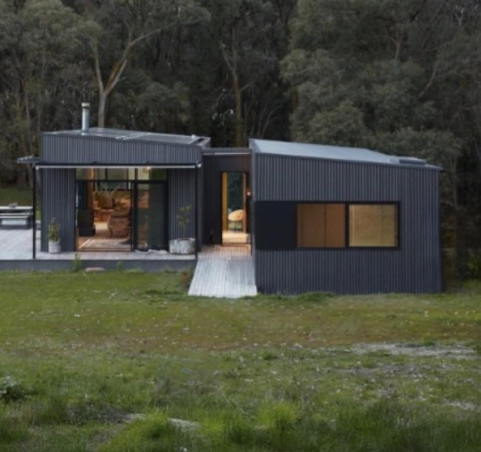Two dark corrugated volumes linked by a timber deck, set in grass and trees