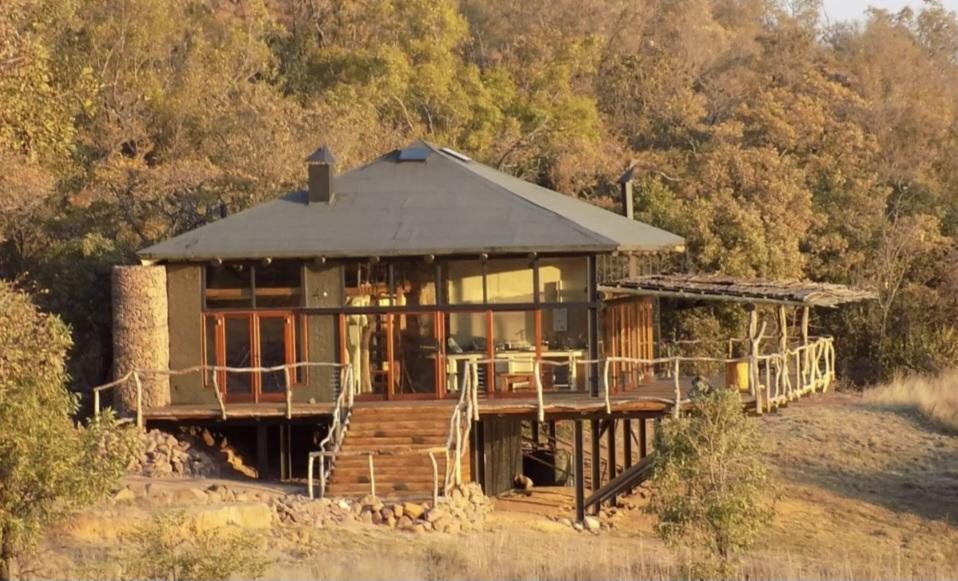 A raised bush lodge with a stone chimney, timber deck and dark roof, set against a wooded slope