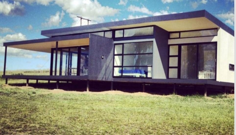 A low glass pavilion on steel stilts in open grassland under a blue sky